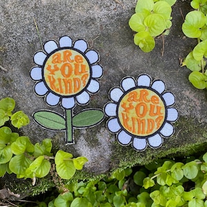 May include: Two flower-shaped patches with the text "Are You Kind?" in orange. The flowers have white petals, yellow centers, and green stems and leaves. The patches are on a textured gray surface, surrounded by green plants.