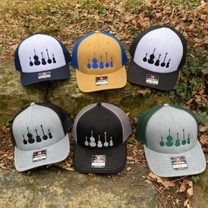 May include: Six baseball caps with a mesh back and a curved brim are displayed. Each cap features a silhouette of stringed instruments. The caps come in various colors, including navy, gold, black, gray, and green.
