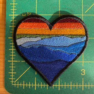 May include: Embroidered heart-shaped patch with a mountain landscape design. The patch features layers of colour, including shades of blue, green, orange, and burgundy. The patch is approximately 7.6 cm wide and has a black border.