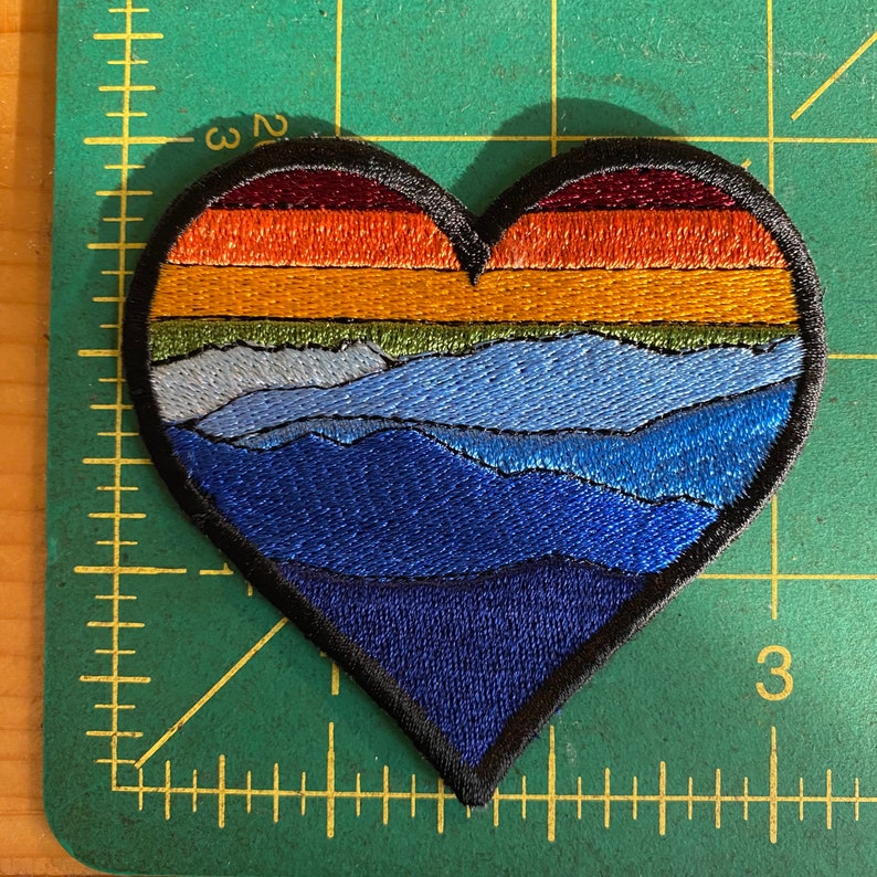 May include: Embroidered heart-shaped patch with a mountain landscape design. The patch features layers of colour, including shades of blue, green, orange, and burgundy. The patch is approximately 7.6 cm wide and has a black border.