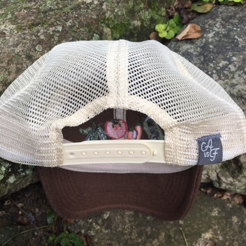 May include: A brown trucker hat with a beige mesh back and a brown brim. The hat features a white adjustable snap closure and a small gray tag with the text "CA vs GF". The interior design includes a tomato and vegetable pattern.