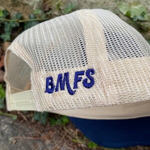 Pode incluir: Bon&eacute; de caminhoneiro bege com parte traseira em malha e aba azul marinho. O bon&eacute; apresenta as letras bordadas "BMFS" em azul marinho na lateral. Fechamento por press&atilde;o ajust&aacute;vel.