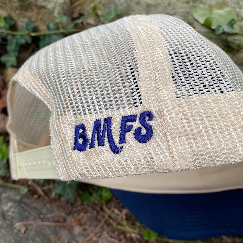 Pode incluir: Bon&eacute; de caminhoneiro bege com parte traseira em malha e aba azul marinho. O bon&eacute; apresenta as letras bordadas "BMFS" em azul marinho na lateral. Fechamento por press&atilde;o ajust&aacute;vel.
