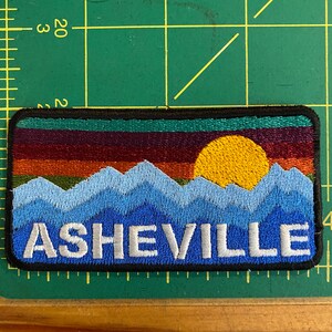 Peut inclure: &Eacute;cusson brod&eacute; repr&eacute;sentant un paysage de montagne avec un coucher de soleil et le mot "ASHEVILLE". Le patch a une bordure noire et est rectangulaire. Les montagnes sont bleues, le coucher de soleil est orange et jaune.