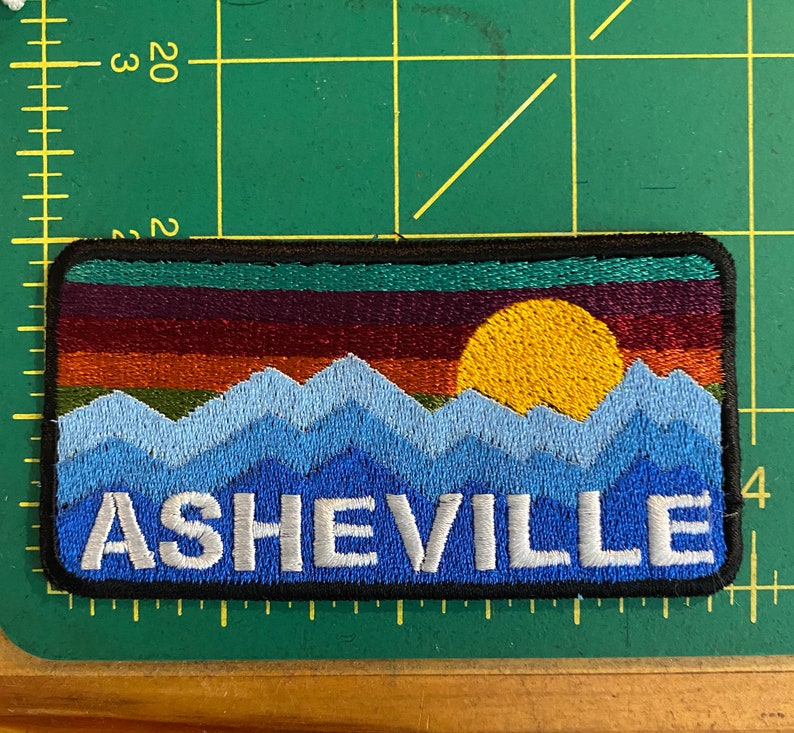 Peut inclure: &Eacute;cusson brod&eacute; repr&eacute;sentant un paysage de montagne avec un coucher de soleil et le mot "ASHEVILLE". Le patch a une bordure noire et est rectangulaire. Les montagnes sont bleues, le coucher de soleil est orange et jaune.