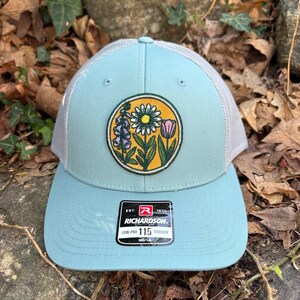 May include: A pale blue trucker hat with a floral patch featuring a daisy, tulip, and other flowers. The hat has a grey mesh back and a curved brim. The patch is circular with a yellow background. The tag reads "RICHARDSON".