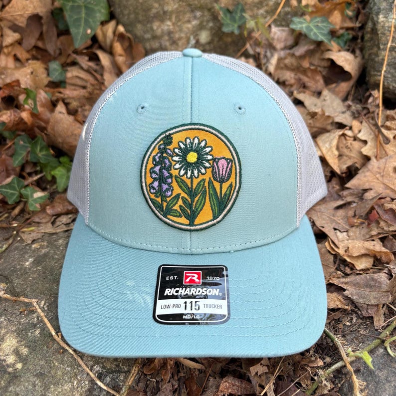 May include: A pale blue trucker hat with a floral patch featuring a daisy, tulip, and other flowers. The hat has a grey mesh back and a curved brim. The patch is circular with a yellow background. The tag reads "RICHARDSON".