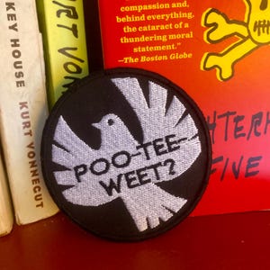 May include: A round, black embroidered patch featuring a white dove design with the text "POO-TEE-WEET?" in white. The patch is set against a backdrop of books with text.
