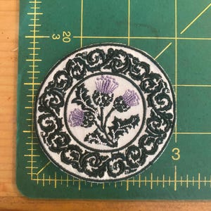 May include: Round embroidered patch featuring a thistle design. The patch has a white center with purple thistles and green leaves. A dark green, ornate border surrounds the central image. The patch is approximately 3 inches in diameter.