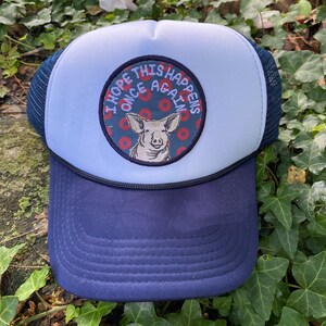 May include: A navy blue and white trucker hat with a patch featuring a pig illustration and the text "I HOPE THIS HAPPENS ONCE AGAIN". The hat has a mesh back and a curved brim.