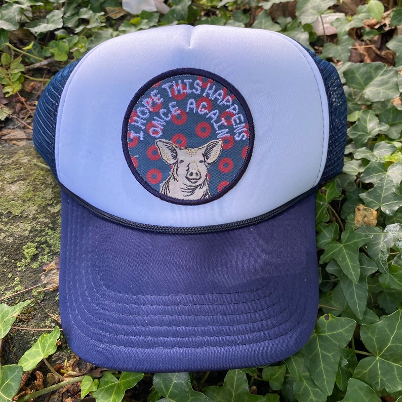 May include: A navy blue and white trucker hat with a patch featuring a pig illustration and the text "I HOPE THIS HAPPENS ONCE AGAIN". The hat has a mesh back and a curved brim.