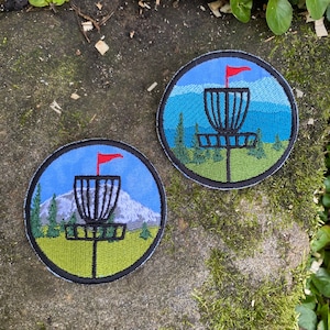 May include: Two round embroidered patches featuring a disc golf basket with a red flag, set against a backdrop of mountains, trees, and a blue sky. The patches have a black border and green and blue color scheme.