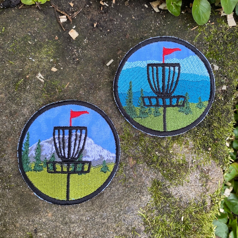 May include: Two circular embroidered patches depicting a disc golf basket with a red flag, set against a backdrop of mountains, trees, and a blue sky. The patches have a black border and a green and blue colour scheme.