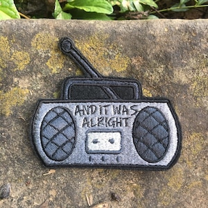 May include: Embroidered patch in the shape of a retro boombox. The patch is primarily gray and black, with the words "AND IT WAS ALRIGHT" embroidered across the front. The boombox has a cassette tape slot and antenna.