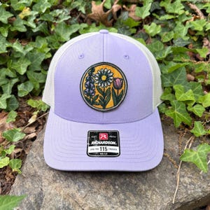 May include: A lilac trucker cap with a white mesh back and a floral patch. The patch features a daisy, tulip, and other flowers in a circular design. The cap has a curved peak and a Richardson tag.