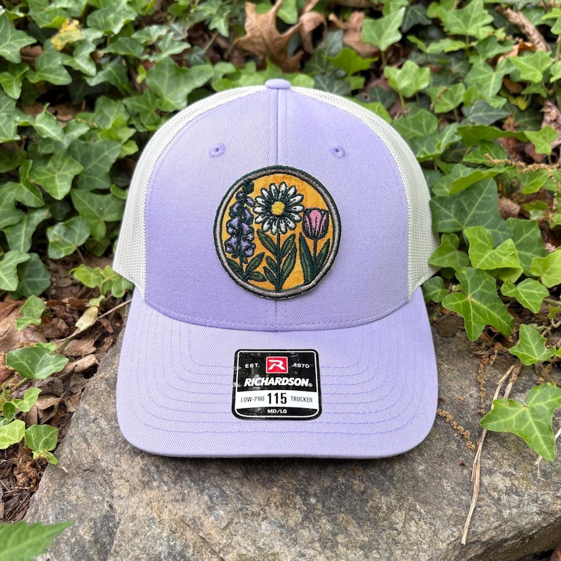 May include: A lilac trucker cap with a white mesh back and a floral patch. The patch features a daisy, tulip, and other flowers in a circular design. The cap has a curved peak and a Richardson tag.