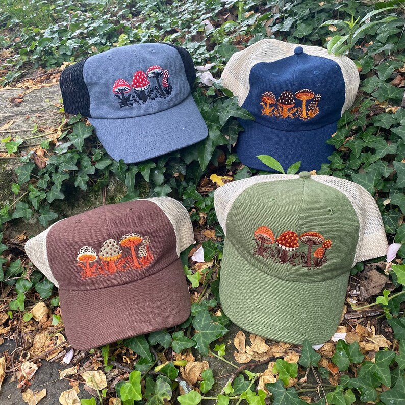 Pode incluir: Quatro bon&eacute;s de caminhoneiro com desenhos de cogumelos bordados. Os bon&eacute;s s&atilde;o em v&aacute;rias cores: azul com uma malha preta, azul marinho com uma malha bege, castanho com uma malha bege e verde com uma malha bege. O bordado de cogumelos apresenta cores vermelhas, laranjas e castanhas.