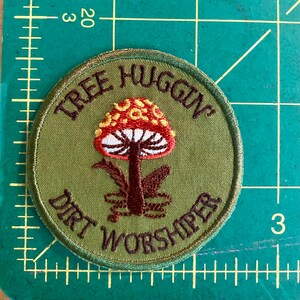 Pu&ograve; includere: Toppa rotonda verde oliva con testo ricamato e un disegno di fungo. Il testo recita "TREE HUGGIN' DIRT WORSHIPER" in filo marrone. Il fungo ha un cappello rosso con macchie gialle e un gambo bianco.