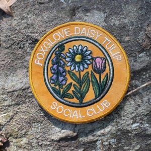 Pu&ograve; includere: Toppa rotonda ricamata con la scritta "FOXGLOVE DAISY TULIP SOCIAL CLUB". La toppa presenta un bordo giallo e illustrazioni di digitale, margherita e tulipano, con foglie e steli verdi, in una scena di giardino.