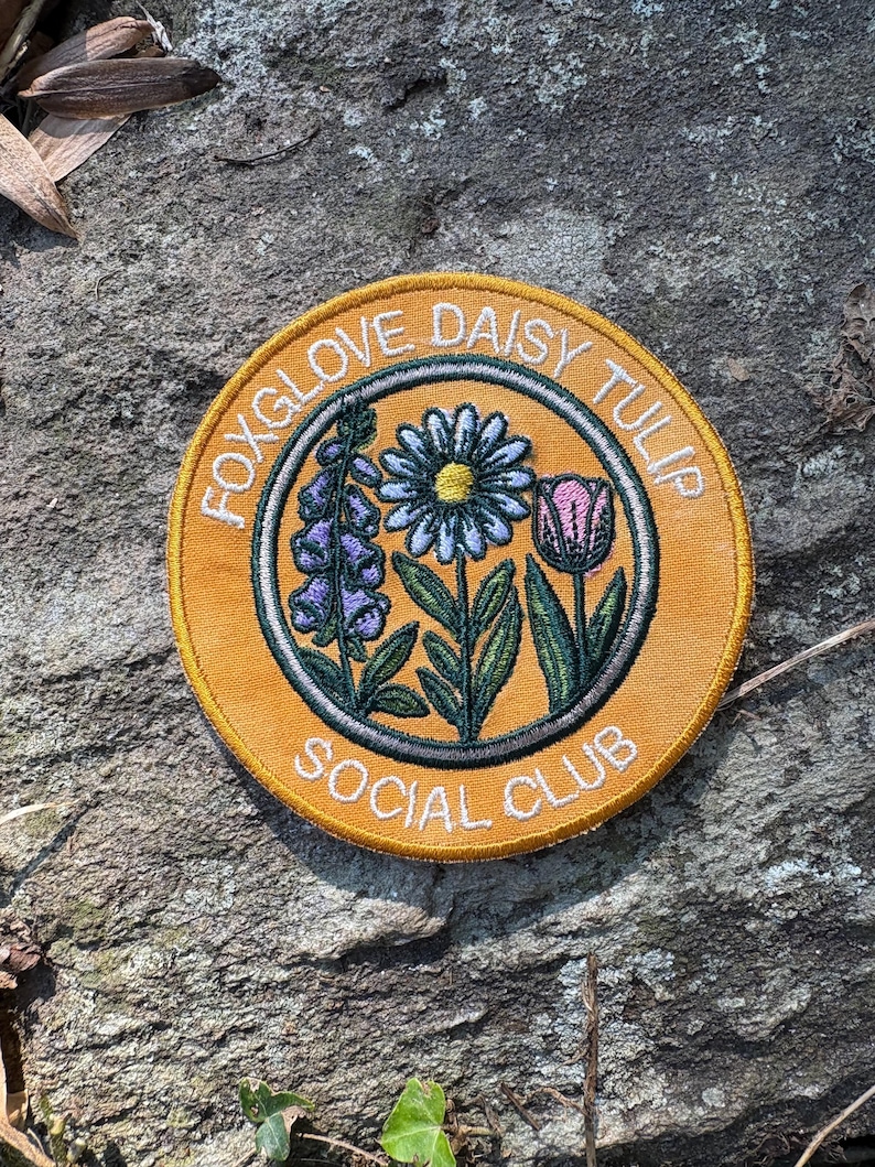Pu&ograve; includere: Toppa rotonda ricamata con la scritta "FOXGLOVE DAISY TULIP SOCIAL CLUB". La toppa presenta un bordo giallo e illustrazioni di digitale, margherita e tulipano, con foglie e steli verdi, in una scena di giardino.