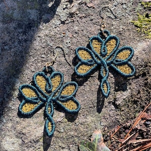 Pode incluir: Um par de brincos em forma de lib&eacute;lula. Os brincos apresentam um contorno verde-azulado escuro com um centro vazado dourado. Os brincos s&atilde;o suspensos por ganchos de orelha prateados. Eles est&atilde;o sobre uma superf&iacute;cie de pedra cinza texturizada.