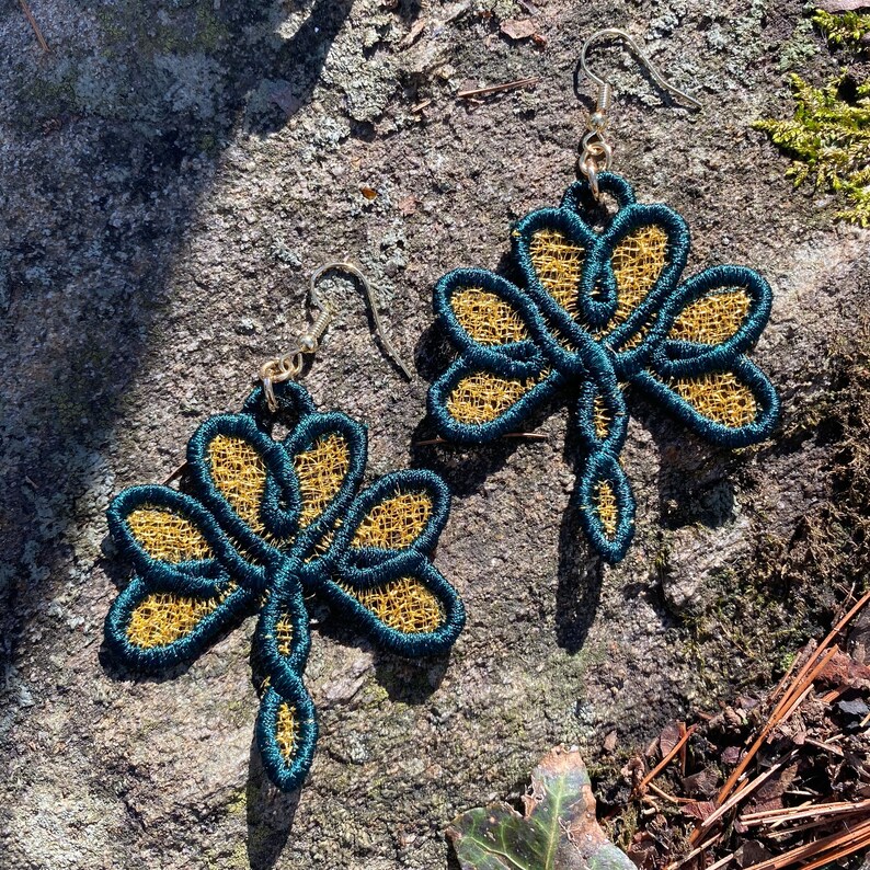 Pode incluir: Um par de brincos em forma de lib&eacute;lula. Os brincos apresentam um contorno verde-azulado escuro com um centro vazado dourado. Os brincos s&atilde;o suspensos por ganchos de orelha prateados. Eles est&atilde;o sobre uma superf&iacute;cie de pedra cinza texturizada.