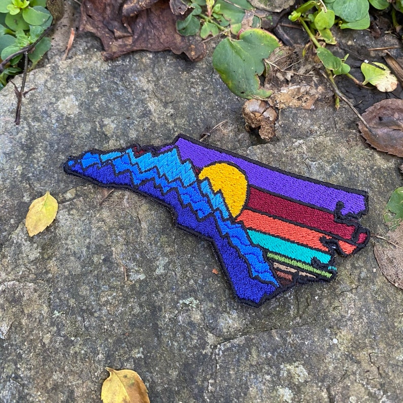 Pode incluir: Remendo bordado em forma de estado, apresentando uma cordilheira, um sol poente e listras coloridas. O remendo &eacute; principalmente azul, roxo, laranja e amarelo, com uma borda preta. O remendo &eacute; provavelmente para uma mochila ou jaqueta.