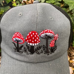 Pode incluir: Bon&eacute; cinza de caminhoneiro com parte traseira em tela preta e aba curva. A frente do bon&eacute; apresenta um design bordado de cogumelos vermelhos com manchas brancas e caules pretos.
