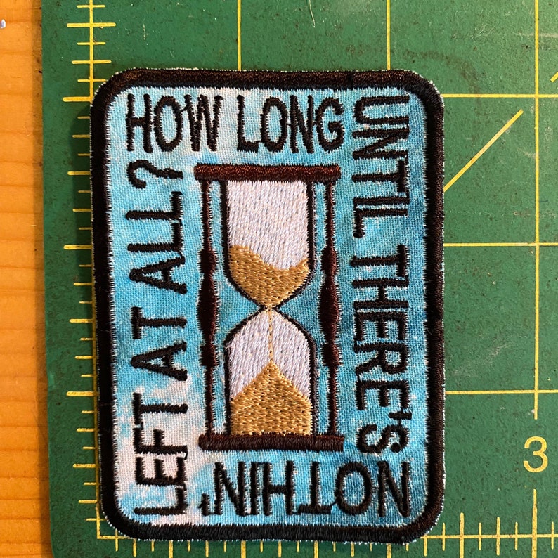 May include: Embroidered patch with a light blue background and a black border. The patch features an hourglass design with the text "HOW LONG LEFT AT ALL? UNTIL THERE'S NOTHING". The hourglass has brown supports and gold sand.