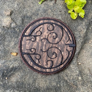 May include: A circular embroidered patch with a brown and black design. The patch features a brown background with a swirling pattern and black embroidered details. The patch is round with a brown border.