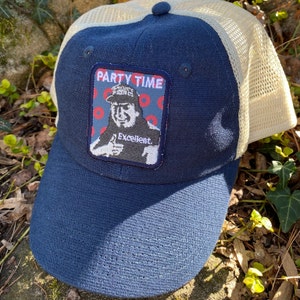 K&ouml;nnte beinhalten: Marineblaue Trucker-Cap mit beigem Mesh-R&uuml;cken. Auf der Vorderseite befindet sich ein Aufn&auml;her mit den Worten "PARTY TIME" und einem Cartoon-Bild eines Mannes, der den Daumen hochh&auml;lt, mit dem Wort "Excellent" darunter.
