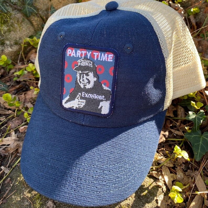 K&ouml;nnte beinhalten: Marineblaue Trucker-Cap mit beigem Mesh-R&uuml;cken. Auf der Vorderseite befindet sich ein Aufn&auml;her mit den Worten "PARTY TIME" und einem Cartoon-Bild eines Mannes, der den Daumen hochh&auml;lt, mit dem Wort "Excellent" darunter.