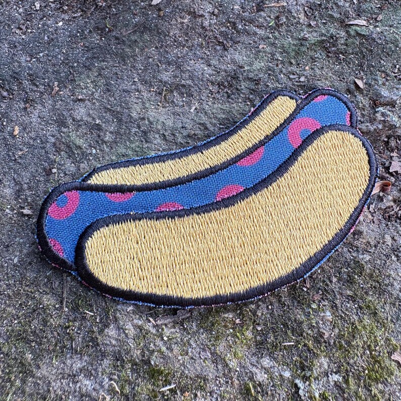 May include: Embroidered patch in the shape of a hot dog. The hot dog bun is yellow with black stitching. The hot dog has a blue and pink polka dot pattern. The patch is on a textured, gray surface.