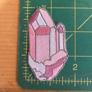 May include: Embroidered patch of a pink crystal cluster. The patch features shades of pink, white, and a silver outline. The crystal design is on a green cutting mat with centimetre markings.