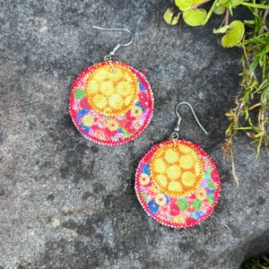 Pu&ograve; includere: Un paio di orecchini in tessuto fatti a mano. Ogni orecchino presenta un vivace motivo floreale colorato su sfondo rosso, con un centro giallo. Gli orecchini hanno ganci per le orecchie color argento.