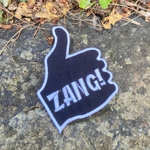 May include: A black and white patch in the shape of a hand giving a thumbs up. The word "ZANG!" is embroidered in white on the patch. The patch has a white border and is lying on a gray rock.