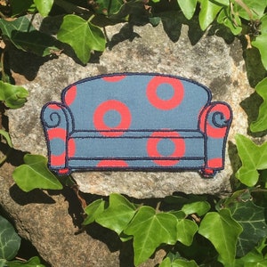 May include: A fabric patch in the shape of a small sofa. The sofa is blue with large red circles and black trim. The patch is set against a stone surface surrounded by green ivy.