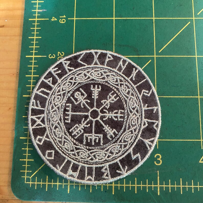 May include: Circular, dark grey patch with a white embroidered Viking compass design. The patch has runes around the edge and detailed knotwork. The patch is likely for clothing or accessories.