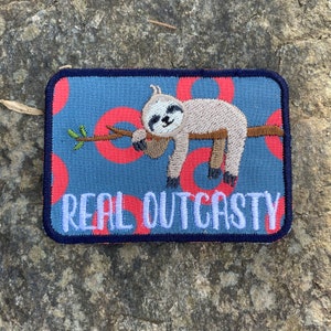 May include: Embroidered patch with a sloth design. The patch shows a sloth on a branch, set against a blue background with red circles. The text "REAL OUTCASTY" is in white, with a navy border.