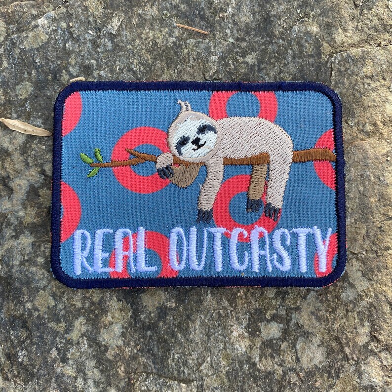 May include: Embroidered patch with a sloth design. The patch shows a sloth on a branch, set against a blue background with red circles. The text "REAL OUTCASTY" is in white, with a navy border.