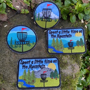 May include: Four embroidered patches with a disc golf theme. Two circular and two rectangular patches feature a disc golf basket, trees, mountains, and the text "Spent a little time on the Mountain". The patches are blue, green, and black.