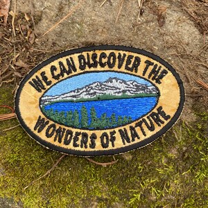 May include: Oval embroidered patch with the text "WE CAN DISCOVER THE WONDERS OF NATURE." The patch features a mountain range, a blue lake, and green trees. The border is black and gold.