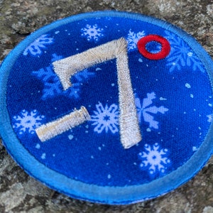 Op de afbeelding: Een ronde, blauw geborduurde patch met een sneeuwvlokpatroon en het nummer 7 in zilveren draad. Er is een kleine rode cirkel aan de bovenkant en de patch heeft een lichtblauwe rand. Geschikt voor kleding of accessoires.