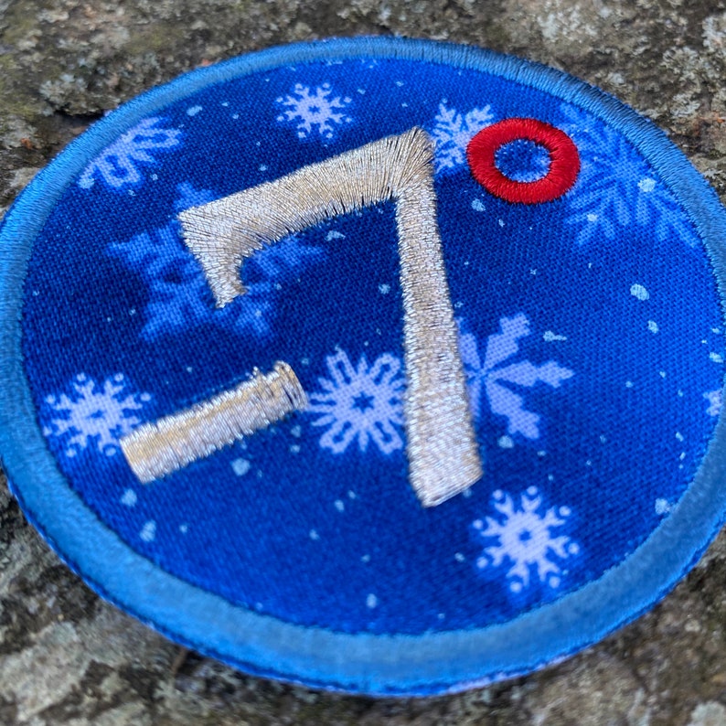 Op de afbeelding: Een ronde, blauw geborduurde patch met een sneeuwvlokpatroon en het nummer 7 in zilveren draad. Er is een kleine rode cirkel aan de bovenkant en de patch heeft een lichtblauwe rand. Geschikt voor kleding of accessoires.