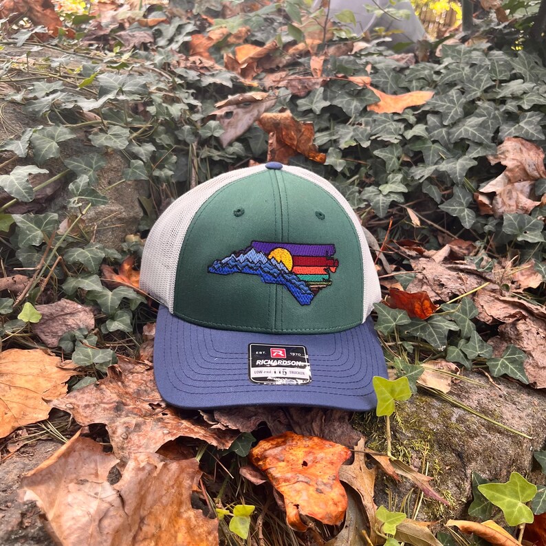 K&ouml;nnte beinhalten: Gr&uuml;n-wei&szlig;e Trucker-M&uuml;tze mit blauem Schirm. Die M&uuml;tze hat ein gesticktes Design der Umrisse des Bundesstaates North Carolina mit einer Bergkette, einem Sonnenuntergang und bunten Streifen.