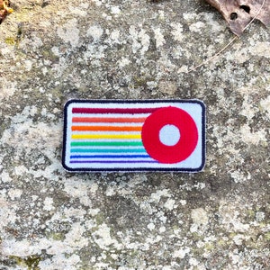 K&ouml;nnte beinhalten: Ein rechteckiger Aufn&auml;her mit einem regenbogenfarbenen Streifendesign und einem roten Kreis mit wei&szlig;em Zentrum. Der Aufn&auml;her hat einen schwarzen Rand und befindet sich auf einem strukturierten grauen Hintergrund.