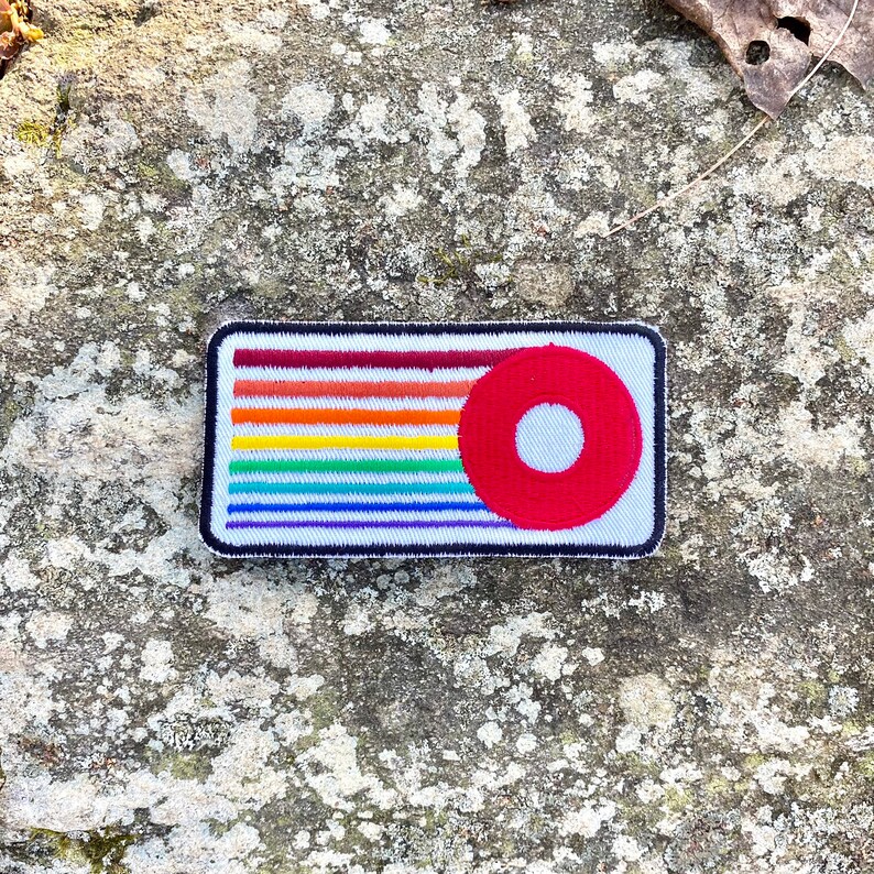 K&ouml;nnte beinhalten: Ein rechteckiger Aufn&auml;her mit einem regenbogenfarbenen Streifendesign und einem roten Kreis mit wei&szlig;em Zentrum. Der Aufn&auml;her hat einen schwarzen Rand und befindet sich auf einem strukturierten grauen Hintergrund.