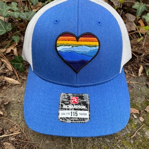 May include: Blue trucker hat with a white mesh back and a heart-shaped patch. The patch features a mountain landscape design in shades of blue, orange, and yellow. The hat has a Richardson 115 Trucker tag.