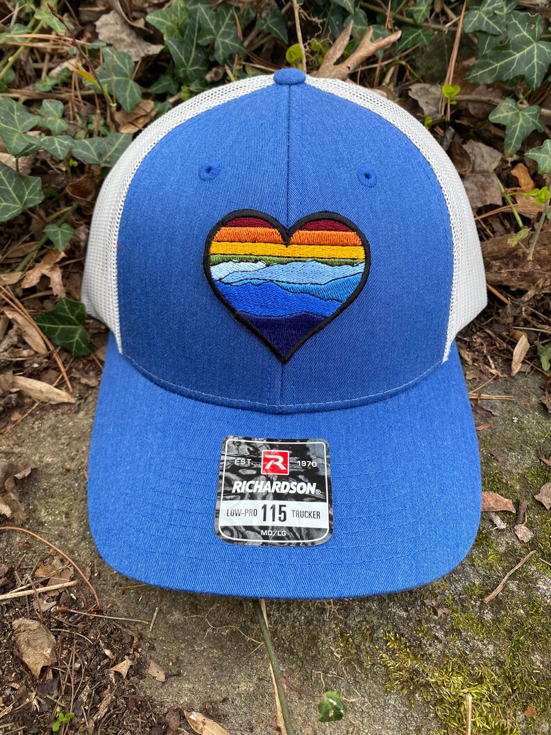 May include: Blue trucker hat with a white mesh back and a heart-shaped patch. The patch features a mountain landscape design in shades of blue, orange, and yellow. The hat has a Richardson 115 Trucker tag.