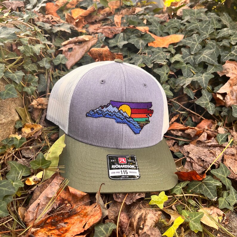K&ouml;nnte beinhalten: Grau-wei&szlig;e Trucker-Cap mit gr&uuml;nem Schirm. Die Kappe zeigt ein gesticktes Design des Umrisses des Bundesstaates North Carolina mit einer Berg- und Sonnenuntergangsgrafik. Die Kappe hat ein Richardson 115 Trucker-Etikett.